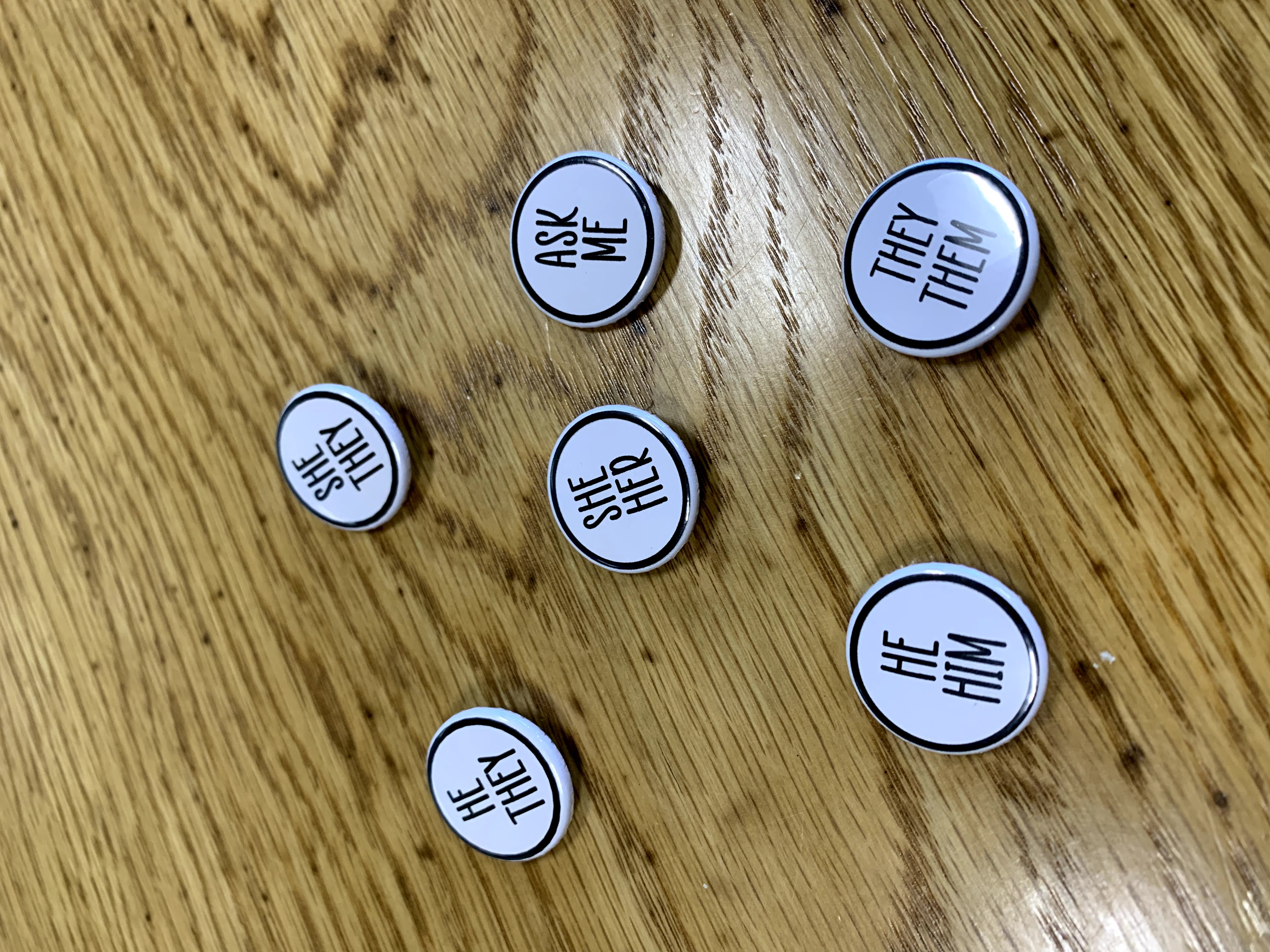 Black and white round buttons on a wood table. The font is all capital letters showing a variety of pronoun pairs. She her, he him, they them, she they, he they, and ask me.