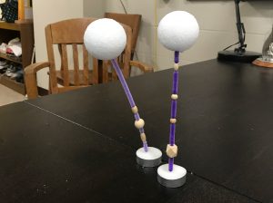 Photograph of two deep-sea mooring models. Mooring models are vertically oriented and constructed with materials purchased at a craft store. A Styrofoam ball at the top to represents the flotation material at the top of the mooring. A pipe cleaner represents the mooring itself with beads on it representing the sensors attached to the mooring. A tea-light at the bottom represents the anchor.