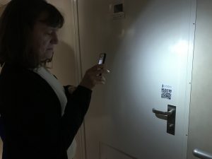 Photo of Amy standing by a door and holding an iPhone in front of her. A QR code label is attached to the door above the handle.