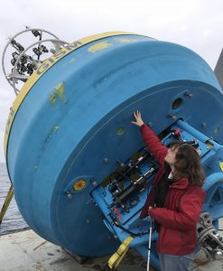 Photo of Amy standing on the back deck reaching to touch the bottom of a very large surface buoy, which is secured leaning over the back edge of the ship. When upright, the total buoy height is about five times as tall as she is.
