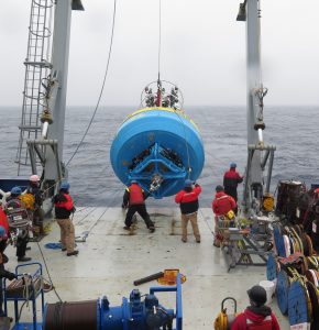 Photo of the surface buoy lifted from the deck and about to go into the water. Several deck crew are helping hold ropes to keep the buoy from swinging. 
